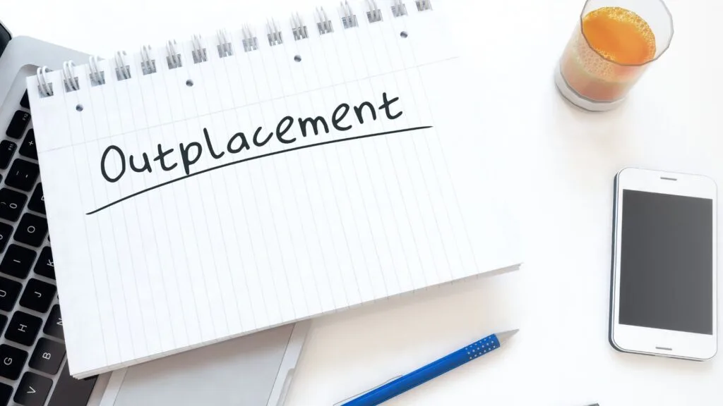 Notitieblok met het woord 'Outplacement' op een bureau met laptop, pen, smartphone en glas sinaasappelsap.