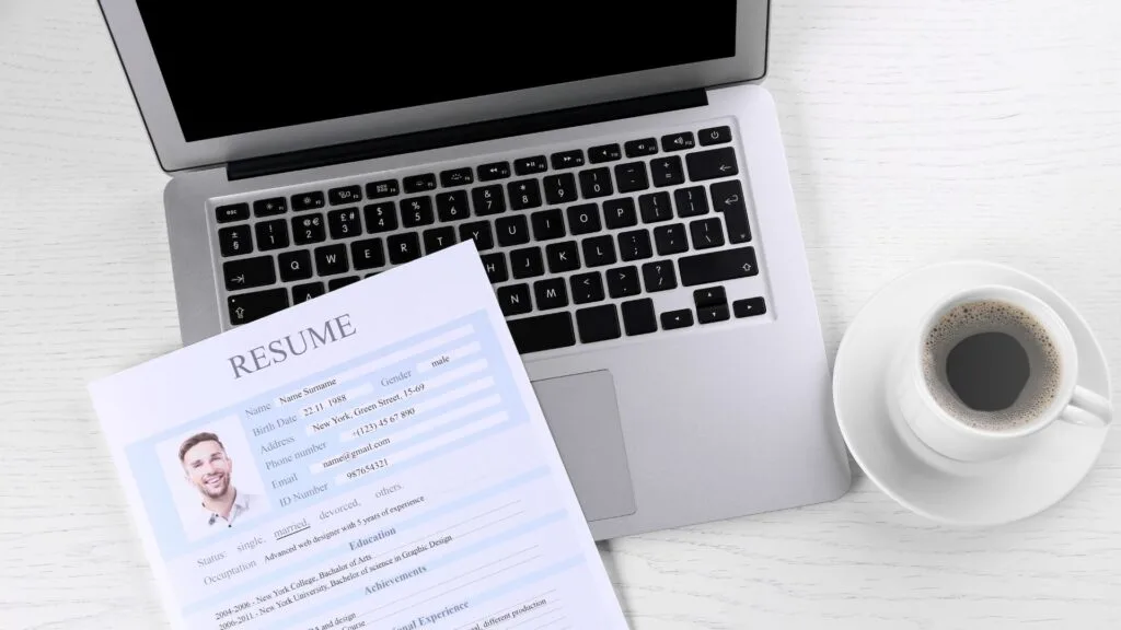 CV met profielfoto ligt op een laptop, met daarnaast een kop koffie op een wit bureau.